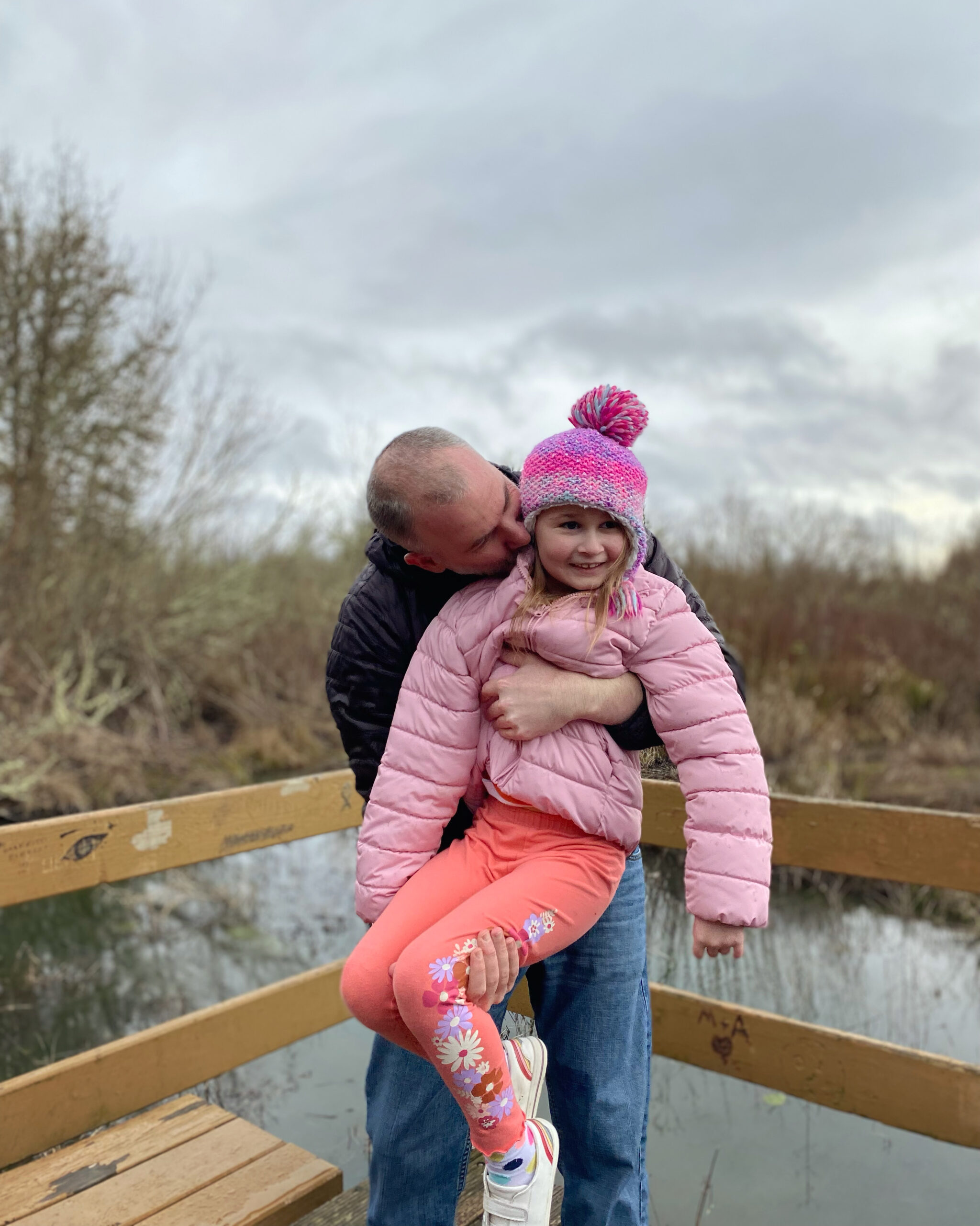 A dad holds up and kisses his six year old blonde daughter on a dock overlooking a marsh in a wetland on a stormy day.