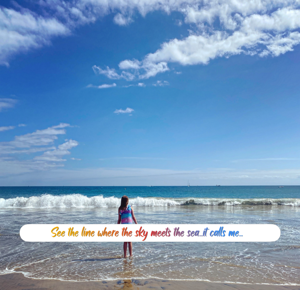 A young girl stands in the sand, watching the waves about to crash at her feet along the shore with a vast blue sky with wispy white clouds above her. The text across the photo reads "See the line where the sky meets the sea, it calls me."