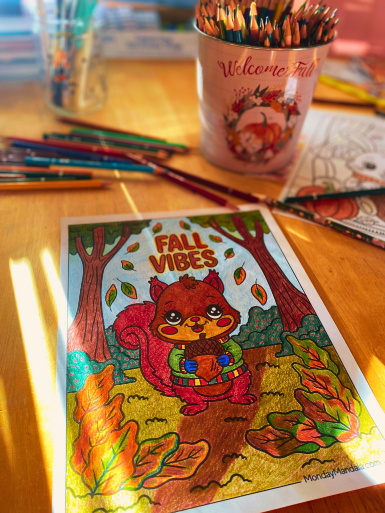 An autumn display decorates a yellow kitchen table. Colored pencils in fall hues surround a coloring page of a cute squirrel in the forest holding a chestnut with the words "Fall Vibes" above his head.